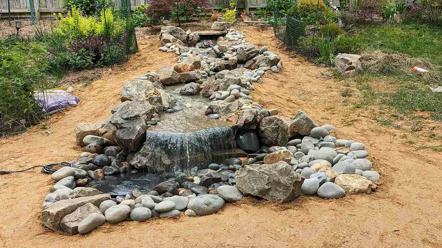 Boulder waterfall feeding a river-rock stream bed through a newly planted garden