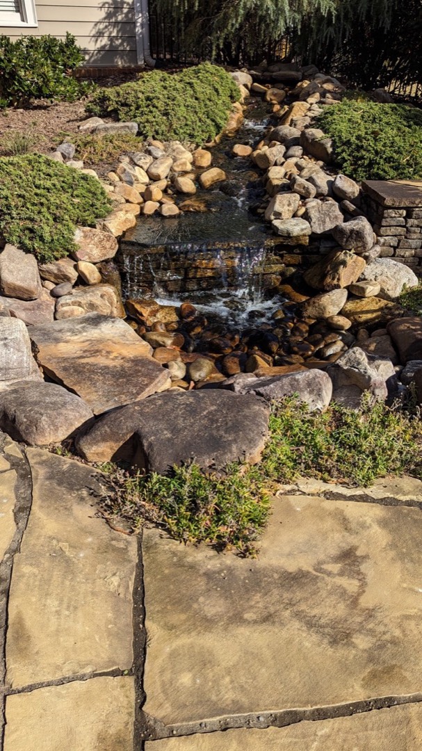 Boulder waterfall and stream spilling down a landscaped slope toward a flagstone patio